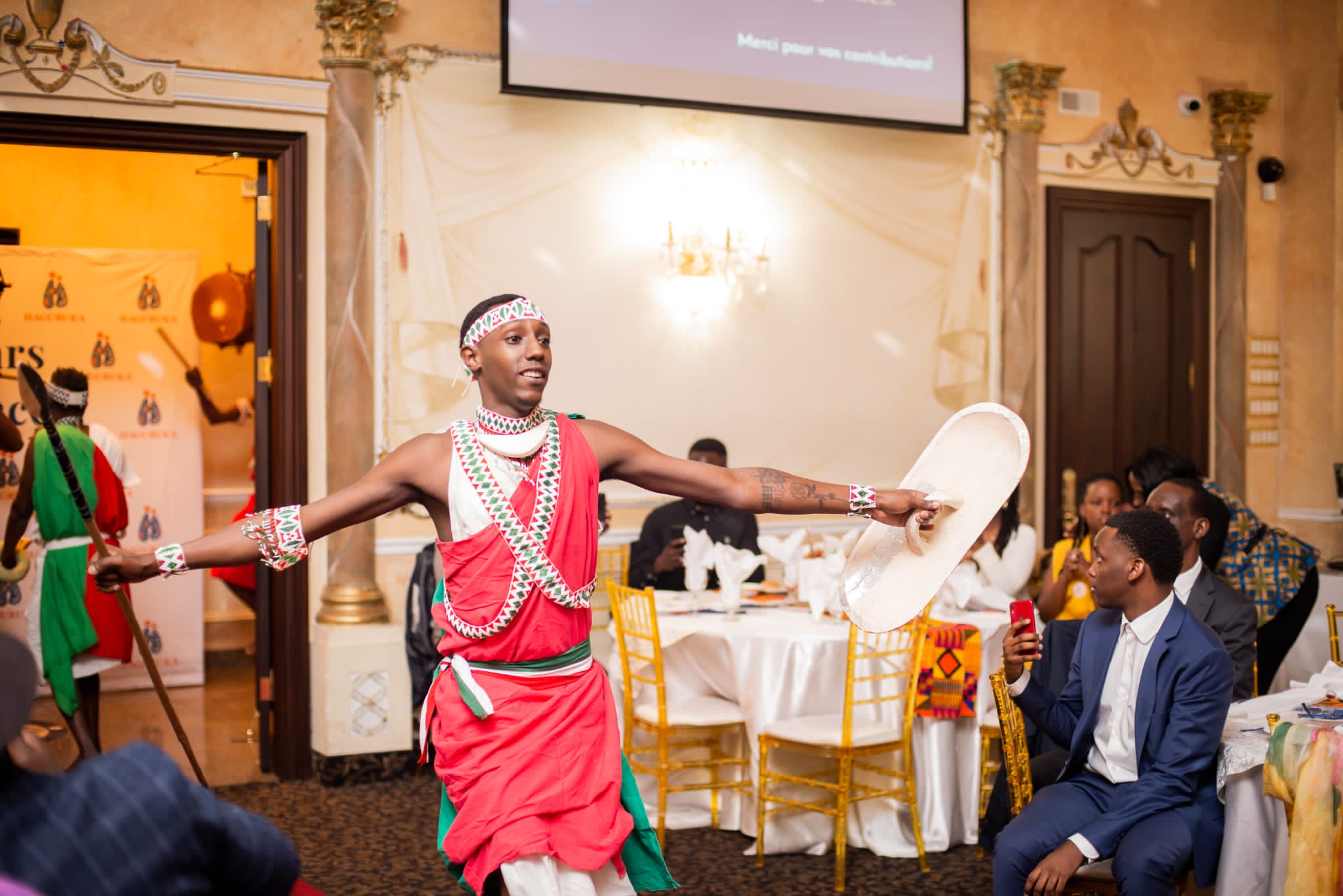A dancer in red regalia sweeps a drum across the floor in front of guests