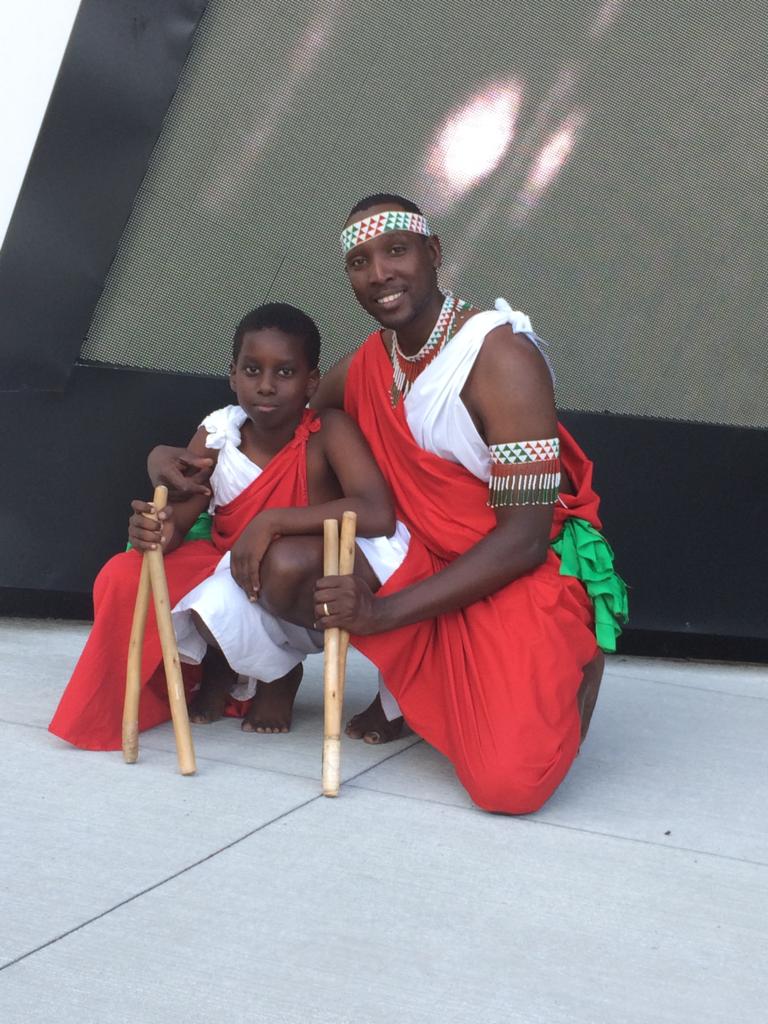 A drummer kneeling beside a child, both in traditional regalia holding drumsticks