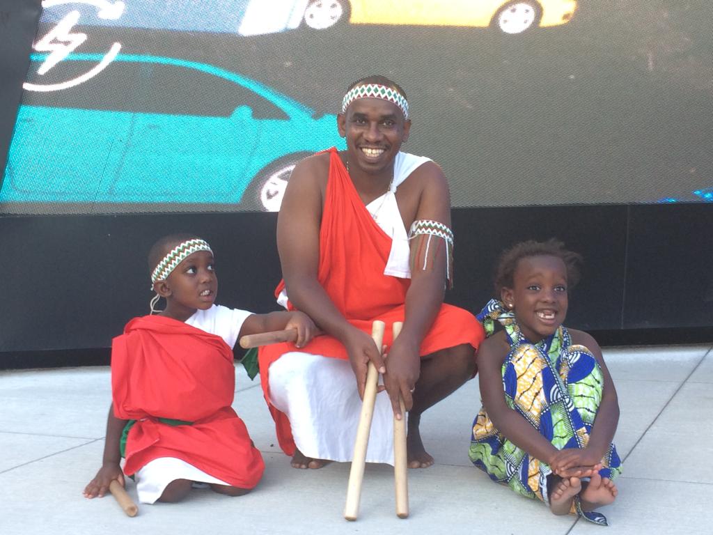 A drummer seated with two children holding drumsticks at a community event